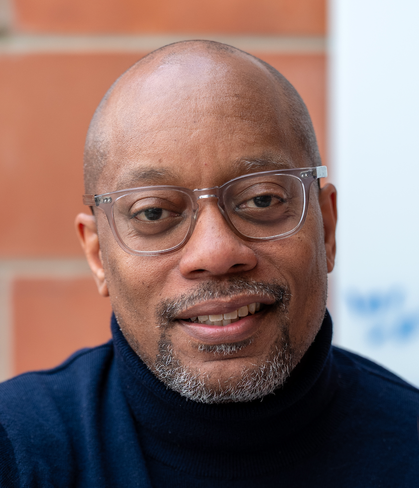 Close-up of a middle-aged black man with a shaved head, graying goatee, and glasses. He is smiling and wearing a navy turtleneck.