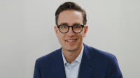 A middle-aged man with short, slicked-back hair, glasses, wearing a button-down light shirt under a navy blazer