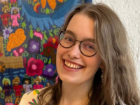 A young woman with long, light brown hair and glasses smiling into the camera against the backdrop of a colorful folk tapestry