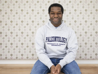 A younger black man with short, natural hair sits in front of a gray-patterned background wearing jeans and a gray UTM Paleontology hoodie.