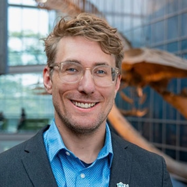 A younger white man with blond, slightly messy short hair and glasses standing smiling in front of a whale skeleton wearing a turquoise button-down underneath a navy suit jacket